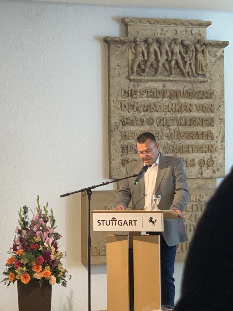 Tomas Brause, Vorsitzender des Gesamtpersonalrats der Landeshauptstadt Stuttgart· bei seiner ansprache am Rednerpult im Stuttgarter Rathaus#auf dem Rednerpult vor ihm das Logo und der Stadt Stuttgart Schriftzug und Pferd. Hinter ihm Gedenktafel