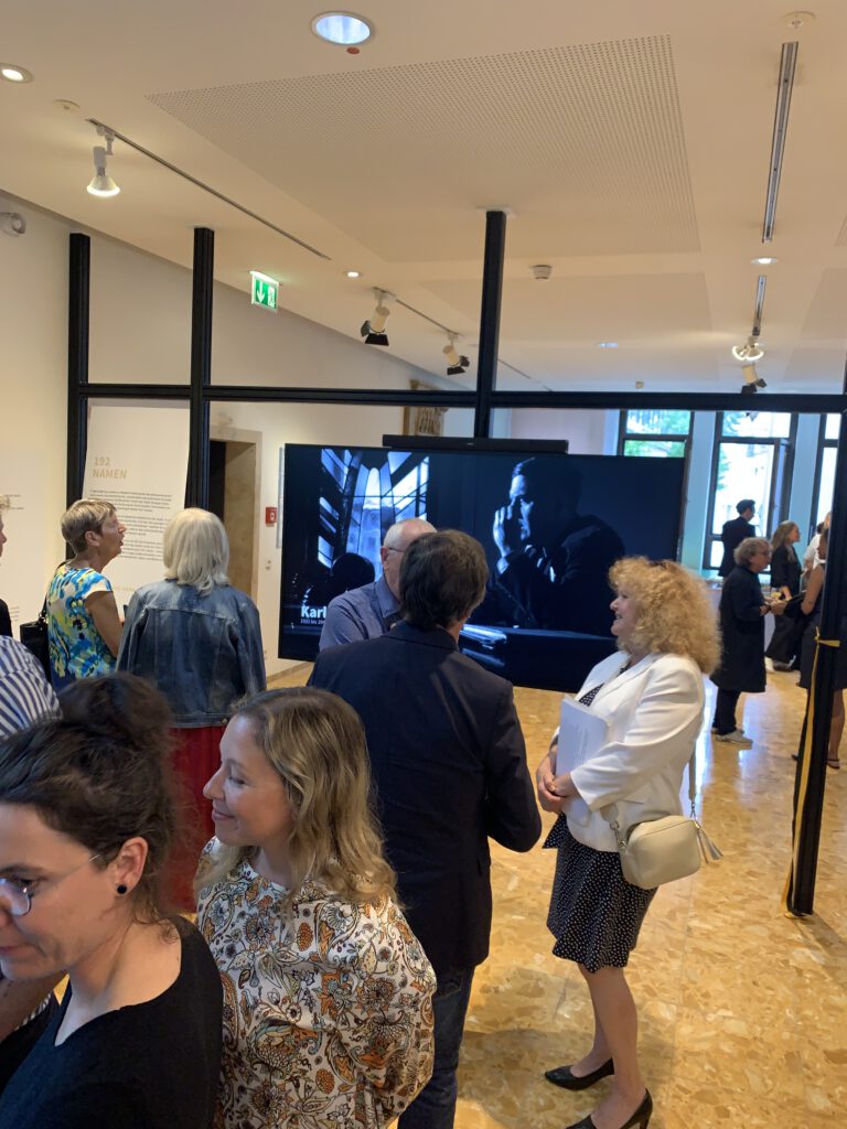 Blick in die Medienstation auf den großen Bildschirm, auf dem der Film" die doppelte Lücke gezeigt wird. RDer Raum ist von einer Gitterstruktur gefasst. Mehrere kleine Gruppen von Ausstellungsbesuchenden im Gespräch. Im Vordergrund Frau Professorin Traub, IRGW .