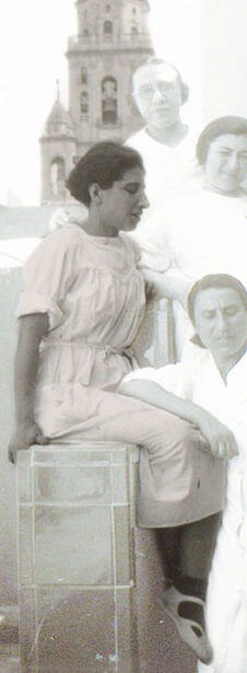 junge Frau in weißem Arbeitshemd im Profil auf der Begrenzung einer steinernen Treppe sitzend rechtsvon ihr absteigend auf der Trreppe posieren ein mann und zwei Frauen in ähnlichen weißen Kitteln. Im Hintergrund ist ein Kirchturmzu sehen. Die Frau ist die junge jüdische Widerstandskämpferin Betty Rosenfeld bei ihrem Arbeitseinsatzals Krankenschwester im spanischen Bürgerkrieg.