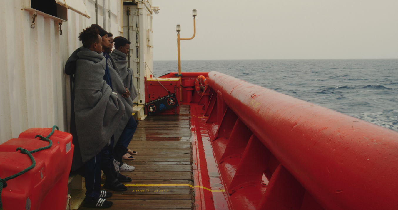 vier Gestalten mit schwarzer Hautfarbe stehen aufgereiht in Decken gewickelt an Deck eines Schiffesund blickenüber die Reling auf das offene Meer