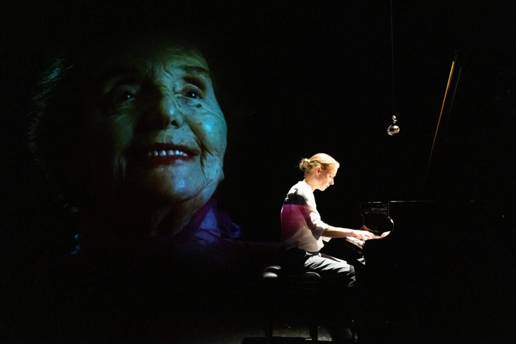 Großaufnahme vom gesicht einer Älteren Dame im dukeln strak für die Bühne geschminkt. Im vordergrund eine mit Spotlicht beleuchtete Frauengestalt, die an einem Konzertflügekl sitzt und spielt