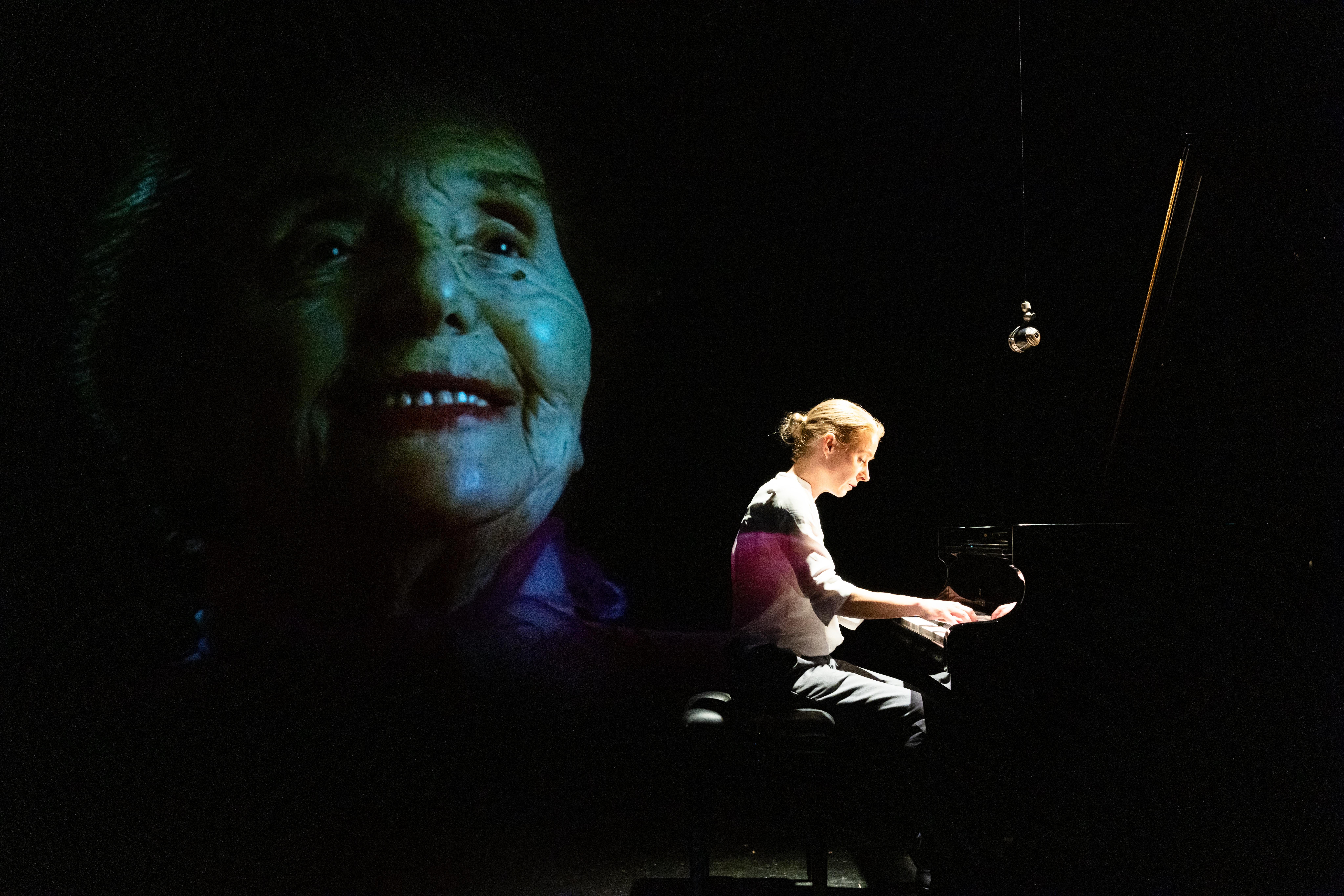 Großaufnahme vom gesicht einer Älteren Dame im dukeln strak für die Bühne geschminkt. Im vordergrund eine mit Spotlicht beleuchtete Frauengestalt, die an einem Konzertflügekl sitzt und spielt