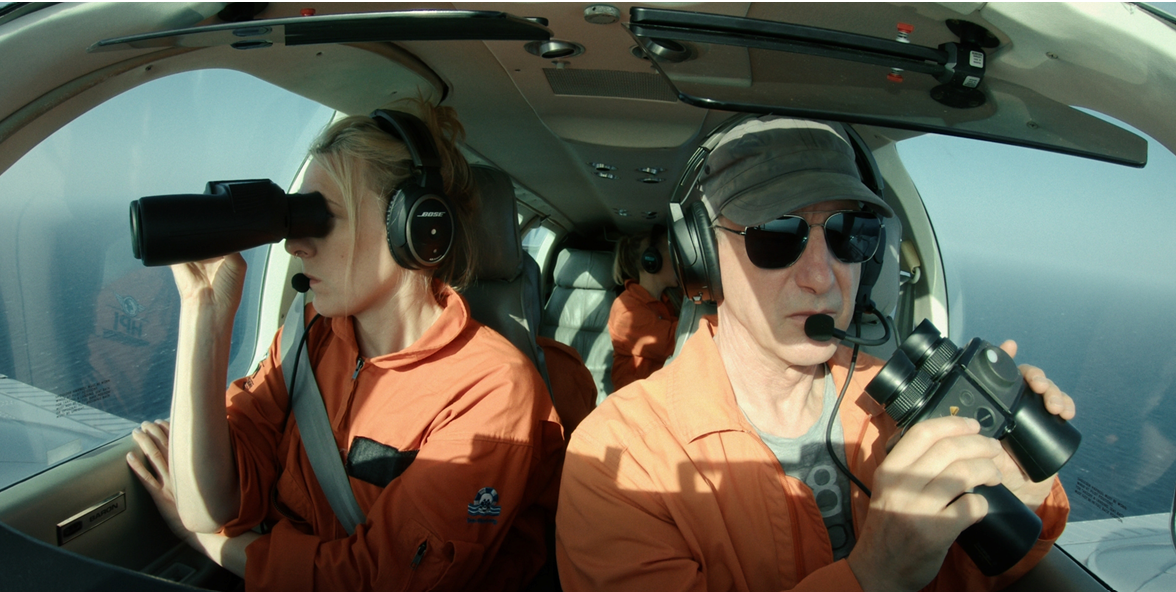 Filmstill aus dem Film kein Land für niemand. zwei personen in orangefarbenen Overalls sitzen in einem Flugzeucockpit. Person links schaut mit dem Fernglas aus dem fenster über das Meer zweite person steuertz das Flugzeug