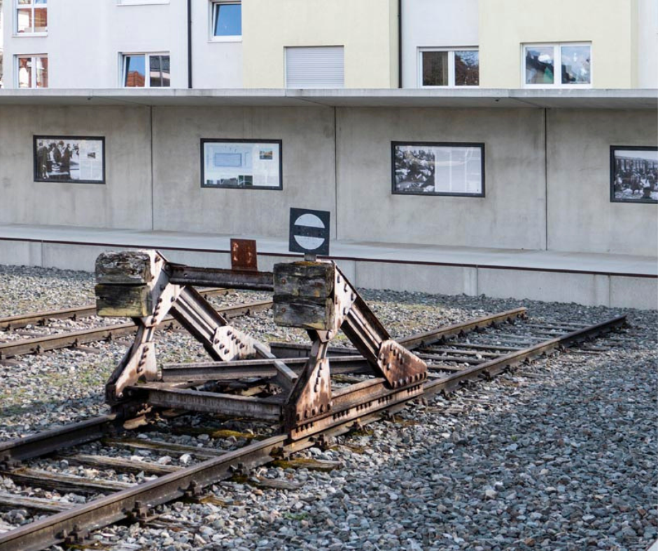 Innenansicht der Gedenkstätte "Zeichen der Erinnerung" Blick über das Gleisbett auf einen historischen Rammbock und die Betonwand mit eingelassenen Ausstellungsschaukästen mit Info zu den Deportationen